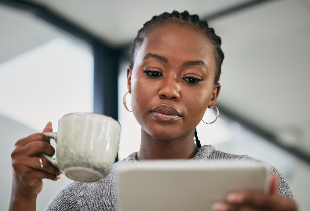 这张照片拍摄的是一位年轻的女商人一边在办公桌上喝咖啡，一边使用数码平板电脑图片下载