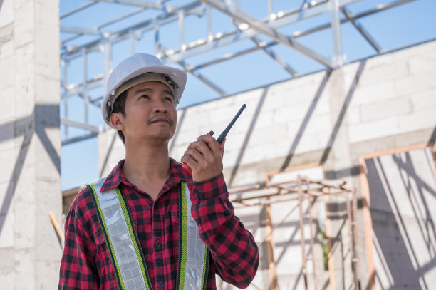 亚洲男子土木工程师施工戴安全白色安全帽站在施工现场手持并使用通信无线电。图片下载
