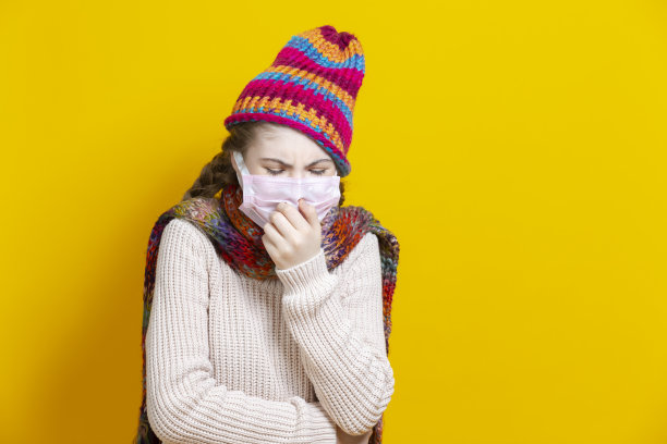 医疗保健的概念。少女在温暖的针织帽子和围巾使用面部防护口罩对抗病毒，而咳嗽时尚的黄色背景。图片下载