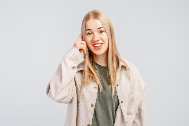 可爱的金发年轻女子说——想想看，把手指放在太阳穴上，穿着休闲服，站在灰色的背景上。肢体语言的概念图片下载