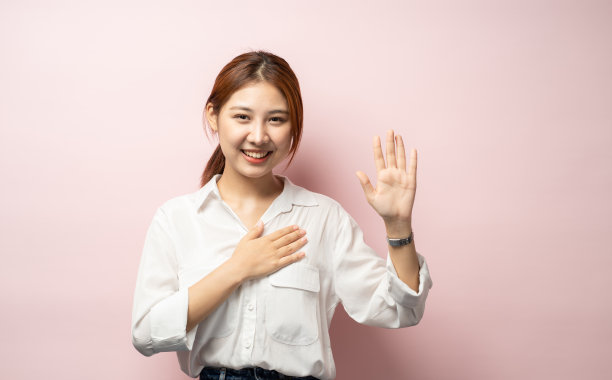 乐观、诚实而可爱的亚洲女孩发誓，发誓说真话，把手放在胸前，举起一只手臂作承诺，表示忠诚和只说真话，站在粉红色的背景。图片下载