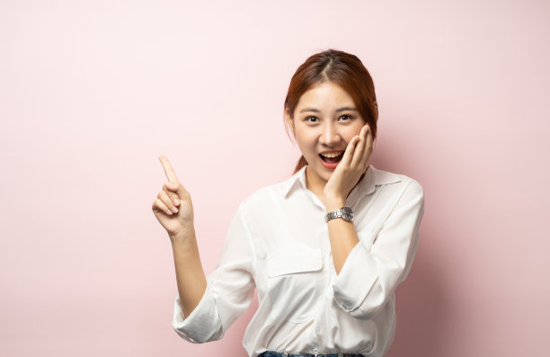 女孩从惊喜和快乐的情绪尖叫，发现商店里有很棒的东西，讲述酷的激动人心的事件，指着左手，盯着相机印象深刻，站在粉红色的背景图片下载