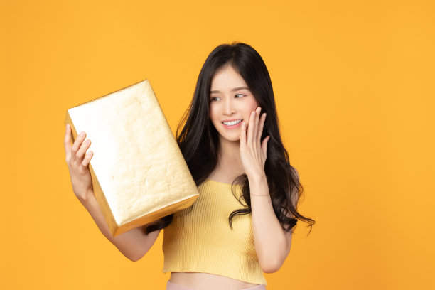 快乐微笑的女孩的肖像在随意地拿着礼品盒和表情脸孤立在黄色背景素材图片