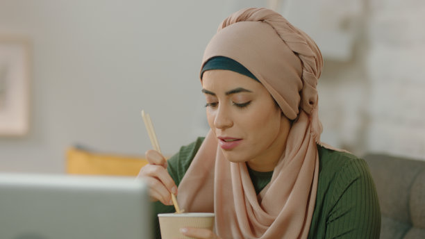 一位戴着头巾的快乐的年轻女子坐在家里的沙发上吃面条，一边看着笔记本电脑上的东西。图片下载