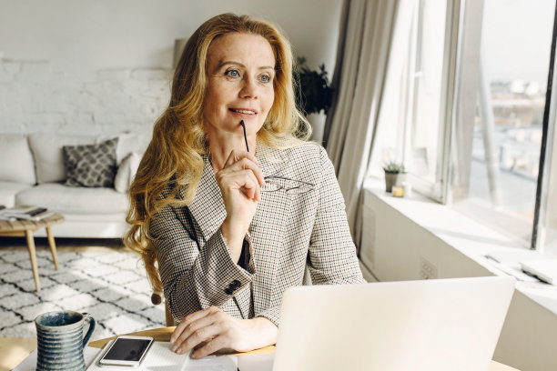 成熟的金发女作家，坐在靠窗的笔记本电脑前，若有所思地凝视着一边，手里拿着眼镜，拿着老式马克杯，桌上有广告文案的手机图片下载