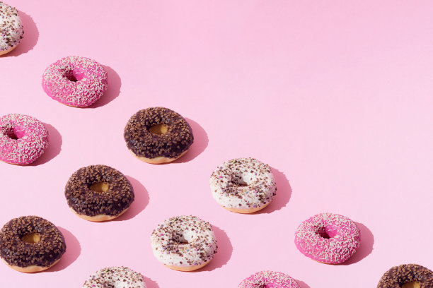 白色、粉色和巧克力色的环形甜甜圈。粉红色粉彩背景上的彩色碎屑。模式。图片下载