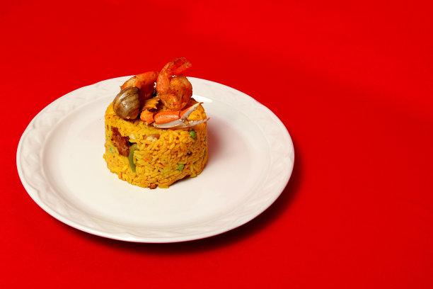 “Arroz a la paella”是西班牙的一种烹饪方法，将藏红花、虾、蛤等海鲜、香肠、西班牙香肠等放在一个白色盘子里，红色背景图片下载
