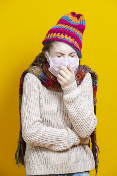 医疗保健的概念。少女在温暖的针织帽子和围巾使用面部防护口罩对抗病毒，而咳嗽时尚的黄色背景。图片下载