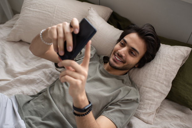 穿着灰色t恤的年轻经理躺在床上用智能手机发短信图片下载