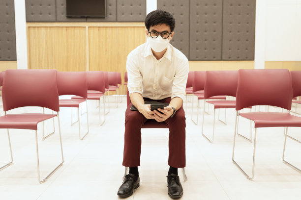 穿着栗红色裤子的亚洲商人，戴着眼镜和面罩，独自坐在医院里等待。图片下载