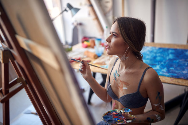 一位穿着胸罩的年轻迷人的女画家正在画室的画架前专注于细节。艺术、绘画、工作室图片下载
