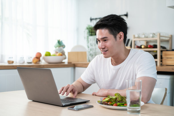 亚洲商人在家工作时吃健康的绿色沙拉。有魅力的帅哥觉得很开心，用笔记本电脑学习在线学习，在厨房吃干净蔬菜保健。图片下载