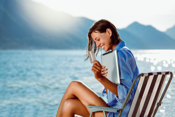 坐在海边，用数码平板电脑和信用卡在线购物的快乐女人。图片下载