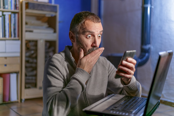 中年男子在笔记本电脑旁边看着自己的手机，露出怀疑的表情。乡村住宅内部与木材燃烧的炉子和堆放的木柴图片下载