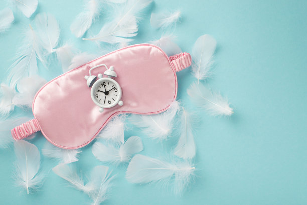 俯视图照片的粉红色丝绸面具睡觉和可爱的小闹钟上的许多白色轻羽毛散落在柔和的天空蓝色孤立的背景copyspace图片下载