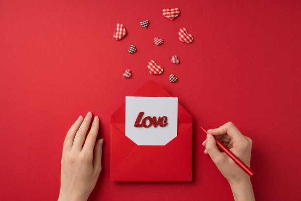 第一人称俯视照片的情人节装饰，女孩的手拿着笔在纸上写着爱在打开的红色信封和格子心孤立的红色背景图片下载