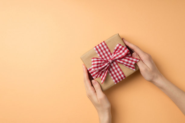 第一人称俯视图照片的情人节装饰女人的手给予工艺纸礼盒与格子丝带蝴蝶结在孤立的米色背景copyspace图片下载