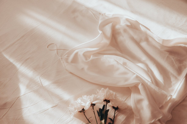 在清晨的阳光下，一件带着细带子的白色丝绸上衣放在一张皱巴巴的床单上，上面还插着一朵菊花图片下载