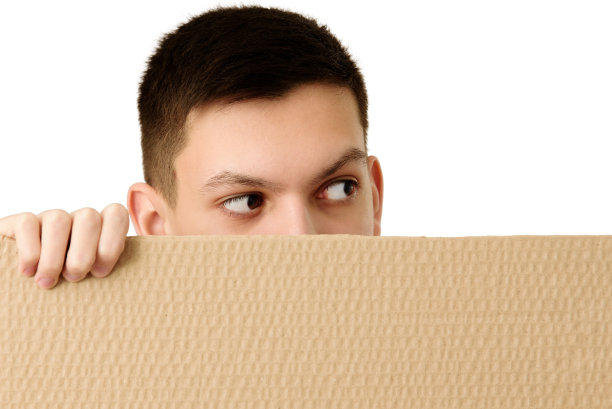 一个十几岁的男孩，在白色的背景上，从空白的棕色纸板横幅后面偷偷地窥视着。特写镜头。副本的空间。图片下载