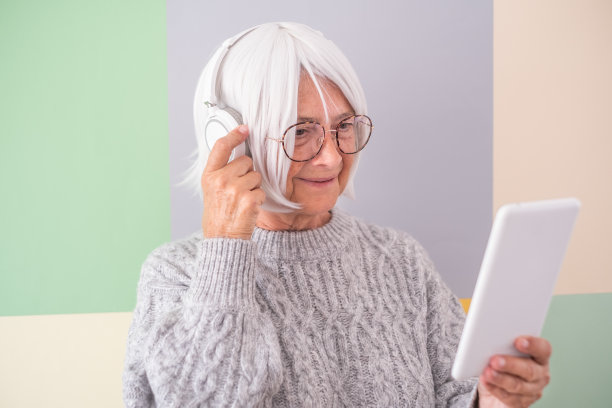吸引人的白人老年妇女拿着数码平板电脑，戴着耳机听音乐，孤立在柔和的颜色背景下，文字的拷贝空间。享受科技和社会的年长女性图片下载