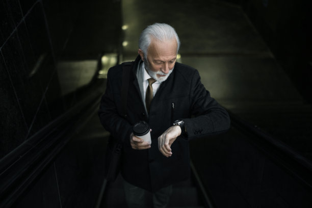 高级商人乘自动扶梯上班，手里拿着一杯咖啡，看着手表图片下载