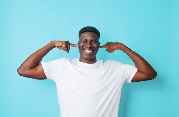 一个帅气的美国人穿着白色t恤，用手指捂住耳朵，用蓝色孤立的背景。图片下载