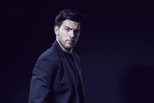 英俊的白人黑发商人穿着黑色西装摆姿势，直视黑色工作室背景图片下载