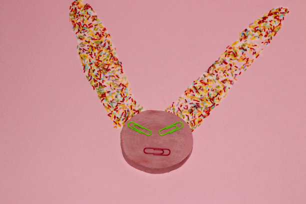 粉红色的木圈，眼睛和嘴巴是用钉书钉做成的，大耳朵是用五颜六色的装饰糖做成的，在粉红色背景的大耳朵创意脸上图片下载