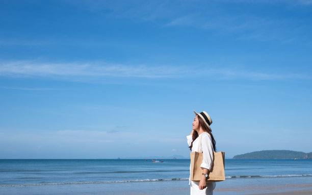 一个美丽的年轻亚洲女人的肖像图像与帽子和包散步在海边与蓝色的天空背景图片下载
