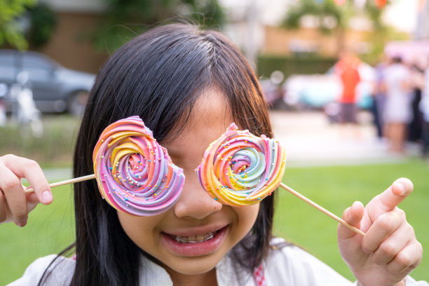 亚洲小女孩幽默地拿着彩虹蛋白糖霜遮住眼睛。图片下载