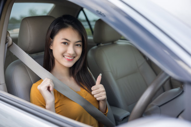 年轻美丽的亚洲妇女得到新车与安全带。她非常高兴和兴奋。她坐下来系上安全带。女司机微笑着开着车走在路上。显示伸出大拇指图片下载