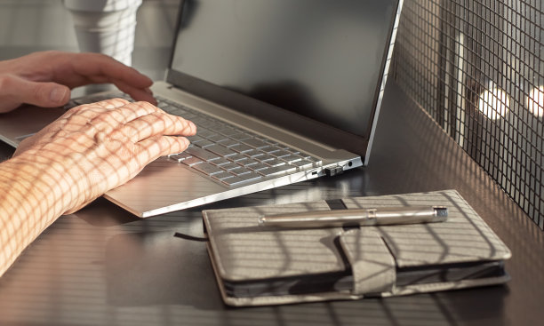 男子的手在笔记本电脑键盘上打字。商人坐在办公室或家庭办公桌前，拿着日记本和硅胶杯，在电脑上写短信或邮件图片下载