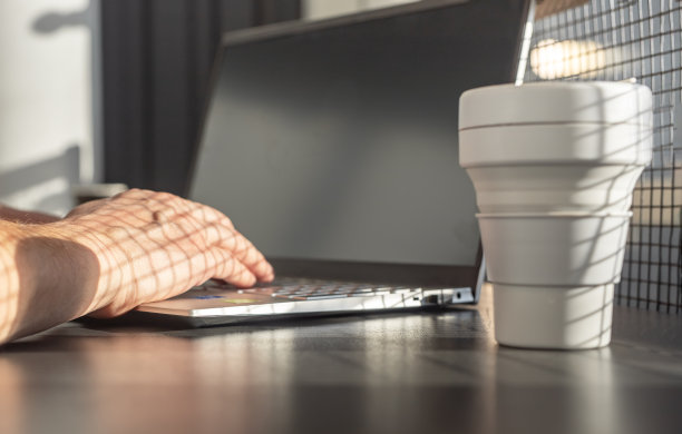 男子的手在笔记本电脑键盘上打字。商人或学生坐在桌旁与电脑和外卖生态硅杯。工作或学习图片下载