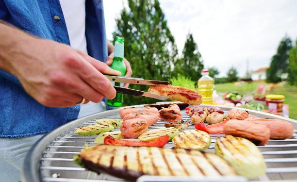 一个男人的手拿着烧烤钳，上面放着美味的油炸香肠，烧烤架上放着香肠、蔬菜、烤西葫芦和胡椒、啤酒瓶、绿色蔬菜和一个公园图片下载