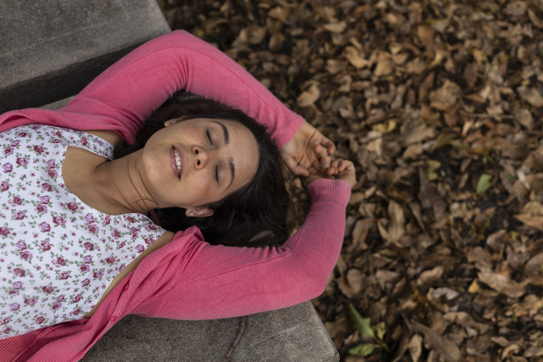 态度积极的年轻的拉丁美洲女性(33岁)对春天的到来感到非常高兴。她仰面躺着，背景是干树叶。她穿着一件粉红色的花裙子。春天的概念。图片下载