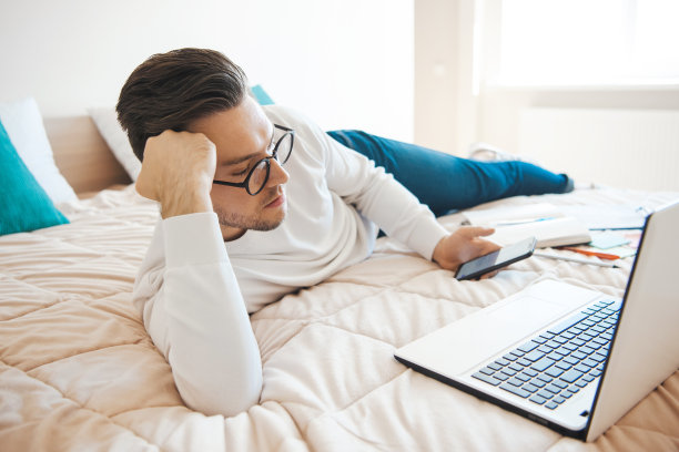 一名白人商人躺在床上，一边用笔记本电脑工作一边用手机打字。人们拿着智能手机。使用电话。图片下载