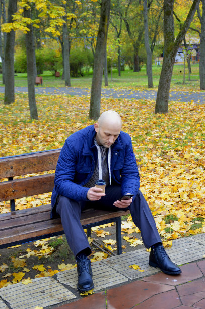 一个男人坐在公园的长椅上，手里拿着一杯咖啡或茶和一部智能手机图片下载