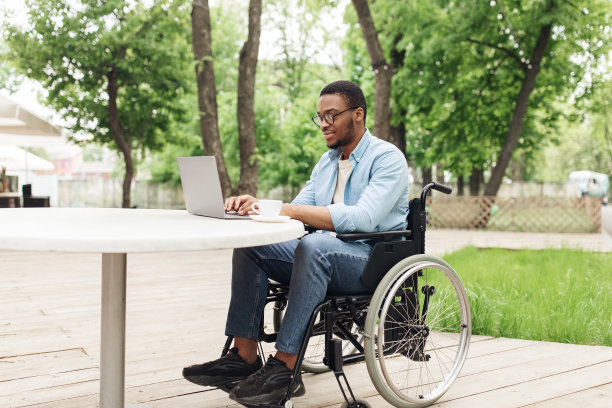 快乐的年轻黑人坐在轮椅上，在公园的咖啡馆用笔记本电脑在线工作，在网上举行商务会议图片下载