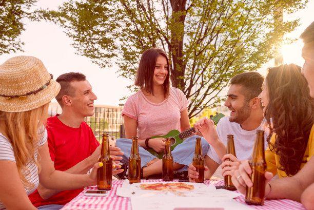 在公园里，年轻人坐在一张铺着桌布、披萨和啤酒的桌子上，一群大学生唱歌、玩耍、一起庆祝，在一个公共公园里举行小型户外派对图片下载