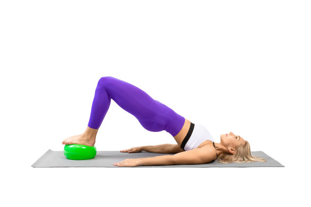 经典的普拉提和道具。女运动员在练习肩桥时，脚下踩着一个小健身球，孤立在白色上。图片下载