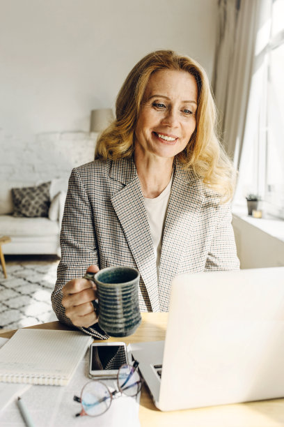 白人中年妇女喝咖啡，与同事用笔记本电脑视频聊天，在休息期间在家工作，智能手机与你的广告的拷贝空间躺在她旁边图片下载