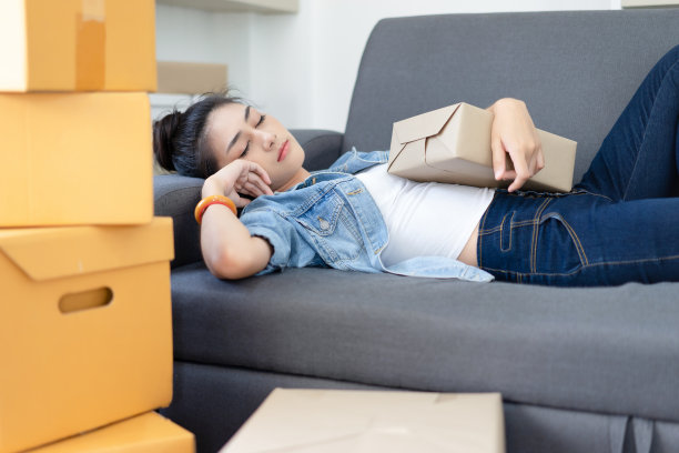 疲惫，年轻的亚洲女性工作了一夜，睡在沙发上与包装盒感到疲惫，白天小睡缺乏能量，失眠或过度工作。超载包装箱。网上销售、电子商务、快递、中小企业图片下载