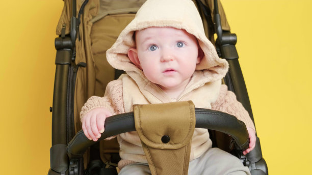 一个快乐的孩子坐在画室黄色背景的婴儿车里。微笑的蹒跚学步的小男孩，在一个变压器婴儿车运送儿童，8个月大图片下载