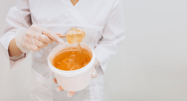 糖脱毛大师将脱毛膏缠绕在刮刀上。打蜡脱毛水疗程序。白色背景的液体黄色糖糊或脱毛用的蜡图片下载