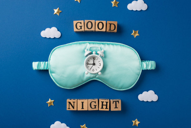 俯视图照片，一个大丝绸浅蓝色睡眠面具和白色的小闹钟，上面散落着星星和云彩形状的五彩纸屑，深蓝色背景上有字母的木制立方体图片下载