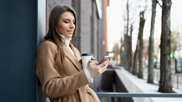 侧面的年轻女子在棕色外套与纸杯咖啡使用智能手机在户外图片下载