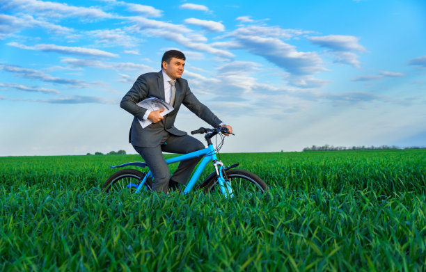 商人穿着西装，骑着一辆带着报告或文件的自行车穿过绿油油的草地，春天的自然美景，经营理念图片下载
