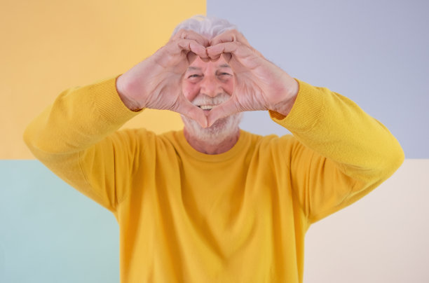面带微笑的老年白人男子与胡子，使心脏形状的手看着相机。英俊的老爷爷站在一个色彩斑斓的孤立的背景上表达爱和感觉图片下载