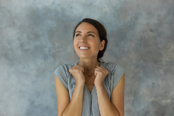 欢快美丽的年轻女子与黑色波浪头发在马尾辫站在灰色和蓝色的墙，向上看一边幸福的微笑紧握拳头与期望中彩票图片下载