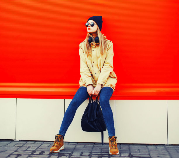 时尚的金发年轻女子模特穿着夹克和背包在城市街道上彩色的红色背景摆姿势图片下载